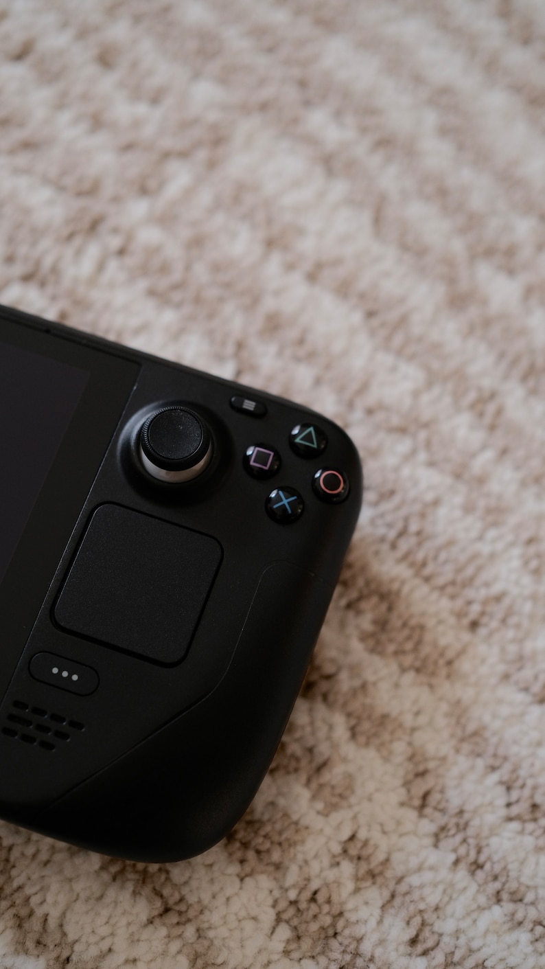 May include: A black handheld gaming device with a joystick, buttons, and a touchpad. The device has a sleek, modern design with a matte finish. The buttons are marked with colored symbols. The device is resting on a light-colored, textured surface.