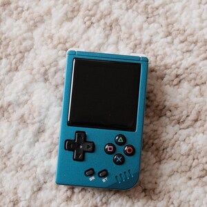 May include: A teal handheld gaming console with a black screen and black control buttons. The console has a directional pad, action buttons, and start/select buttons. The device is resting on a textured, off-white surface.