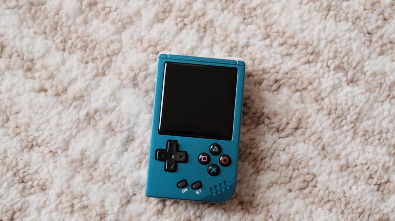 May include: A teal handheld gaming console with a black screen and black control buttons. The console has a directional pad, action buttons, and start/select buttons. The device is resting on a textured, off-white surface.