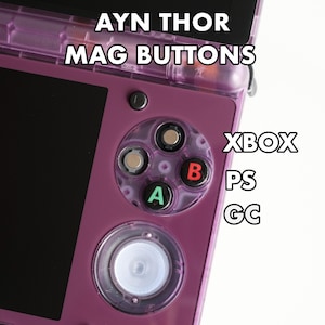 Può includere: Un dispositivo di gioco portatile viola con il testo "AYN THOR MAG BUTTONS". Il dispositivo ha uno schermo nero e pulsanti colorati con le etichette A, B e un pad direzionale. È visibile anche il testo "XBOX, PS, GC".