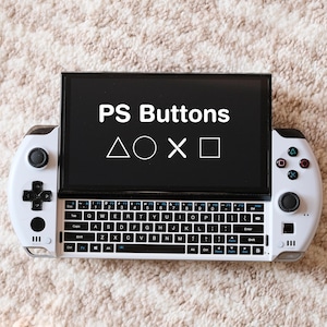 Może przedstawiać: Biała przenośna konsola do gier z czarnym ekranem wyświetlającym "PS Buttons" i symbole PlayStation. Konsola posiada pełną klawiaturę i przyciski kontrolera po obu stronach. Urządzenie znajduje się na teksturowanej, jasnej powierzchni.