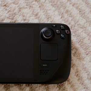 May include: A black handheld gaming console with a large screen and integrated controls. The device features a joystick, buttons, and a touchpad. The screen is off, displaying a black surface. The console is resting on a textured, light-colored surface.