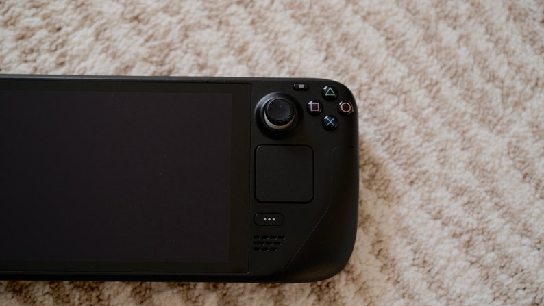 May include: A black handheld gaming console with a large screen and integrated controls. The device features a joystick, buttons, and a touchpad. The screen is off, displaying a black surface. The console is resting on a textured, light-colored surface.