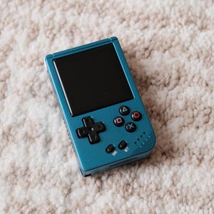 May include: Teal handheld gaming console with a black screen, directional pad, and colored action buttons. The console has a rectangular shape and is resting on a textured, off-white and brown surface.