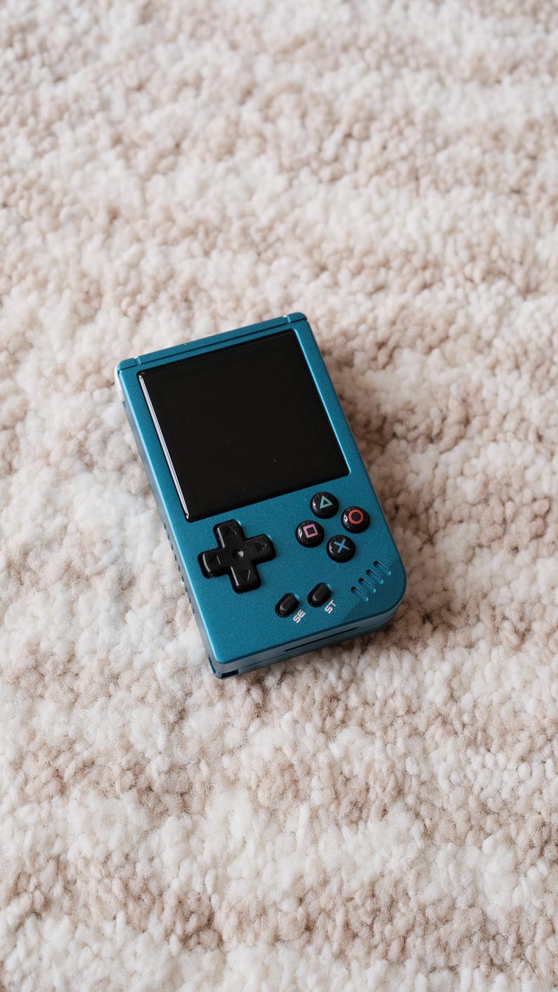May include: Teal handheld gaming console with a black screen, directional pad, and colored action buttons. The console has a rectangular shape and is resting on a textured, off-white and brown surface.