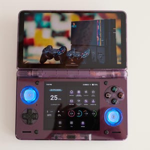 May include: A purple handheld gaming device with a screen displaying a console and controllers. The device has blue illuminated buttons and a screen displaying game information. The device is open and resting on a white surface.