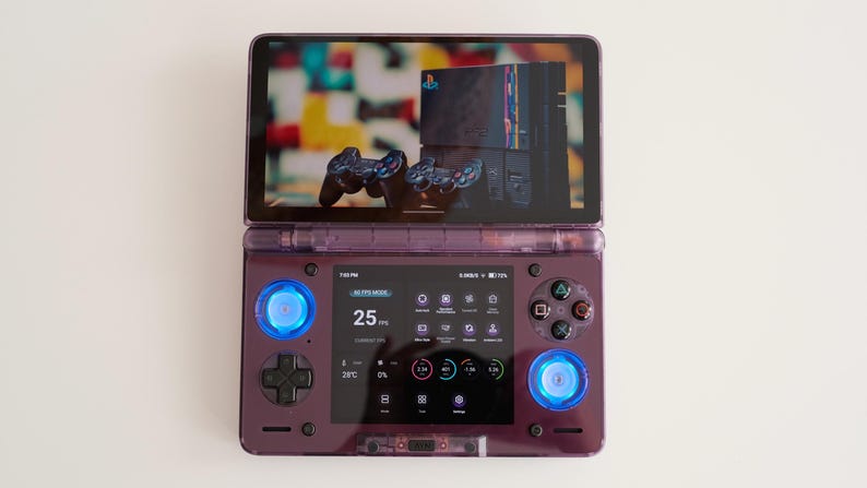 May include: A purple handheld gaming device with a screen displaying a console and controllers. The device has blue illuminated buttons and a screen displaying game information. The device is open and resting on a white surface.