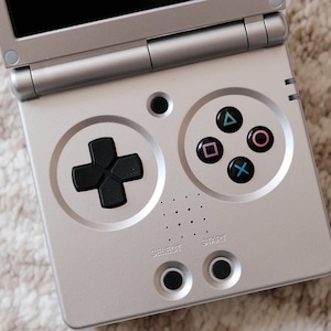 Op de afbeelding: Een zilveren handheld-gameconsole met een zwarte directionele pad en vier face-knoppen. De knoppen zijn gelabeld met de symbolen: driehoek, cirkel, vierkant en X. De console heeft ook een select-knop en een start-knop.