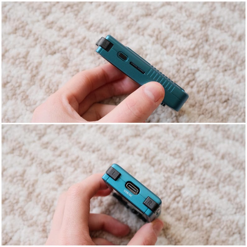 May include: A teal handheld gaming device with a black strap and buttons. The device has a USB/DC port and an SD card slot. The device is held in a person's hand.