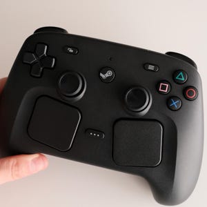Może przedstawiać: Matowy czarny kontroler do gier wideo z centralnym przyciskiem z logo Steam, dwoma analogowymi drążkami, padem kierunkowym oraz czterema przyciskami akcji oznaczonymi niebieskimi, czerwonymi, r&oacute;żowymi i zielonymi symbolami oraz dwoma dużymi touchpadami.