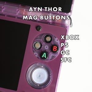 May include: A close-up of a purple handheld gaming device with the text "AYN THOR MAG BUTTONS." The device has a black screen and a set of coloured buttons labelled A, B, and a directional pad. The text "XBOX PS GC SFC" is also visible.