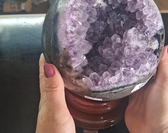 May include: A purple amethyst geode sphere on a wooden stand. The geode is hollow and filled with crystals.