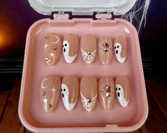 May include: A set of ten almond-shaped press-on nails in a light pink plastic case. The nails feature a nude base with white designs including moons, stars, ghosts, and spider webs, accented with small silver rhinestones on several nails.
