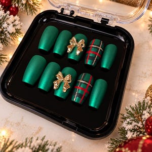 May include: A set of ten press-on nails in a black display case. The nails feature a matte forest green finish, with two nails adorned with gold-toned metallic bows and two nails featuring a red, black, and gold glitter plaid pattern.
