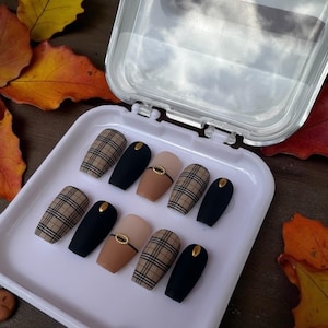 May include: A set of press-on nails in a white plastic case. The nails feature a variety of designs, including a plaid pattern, solid black, and a nude color with a gold accent. The nails are oval-shaped.