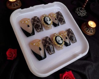 May include: A set of press-on nails with a gothic theme. The nails feature a black and nude color scheme, with intricate damask patterns and cameo accents. Some nails have a black French tip design, and others are adorned with gold and pearl embellishments.