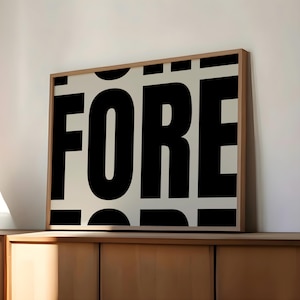 May include: Framed art print with the word "FORE" in large, bold, black letters. The letters are set against a cream-colored background. The artwork is in a wooden frame and rests on a wooden cabinet.