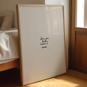 Może przedstawiać: Oprawiony wydruk z ramą z jasnego drewna. Wydruk zawiera ręcznie napisaną frazę "love you to the moon & back" czarnym pismem na białym tle. Rama opiera się o ścianę w pobliżu łóżka.