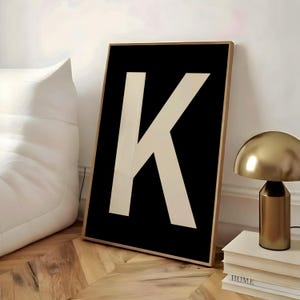 May include: A framed art print featuring a large, cream-colored letter "K" against a black background. The artwork is in a gold-colored frame and leans against a white wall. A gold-colored lamp and books are visible.
