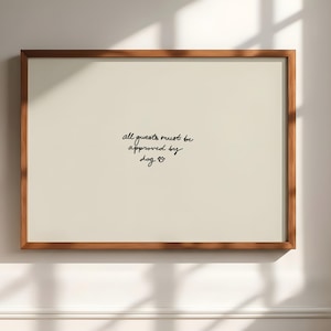 Puede incluir: Una impresión enmarcada con un fondo beige claro y un marco de madera marrón. El texto "all guests must be approved by dog" está escrito en escritura negra. Una huella de pata de perro está al final del texto.