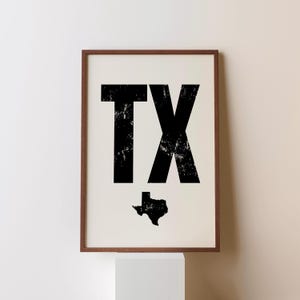 May include: A framed print featuring the letters "TX" in bold, distressed black font, with a black silhouette of the state of Texas below. The print has a cream-colored background and a brown wooden frame.