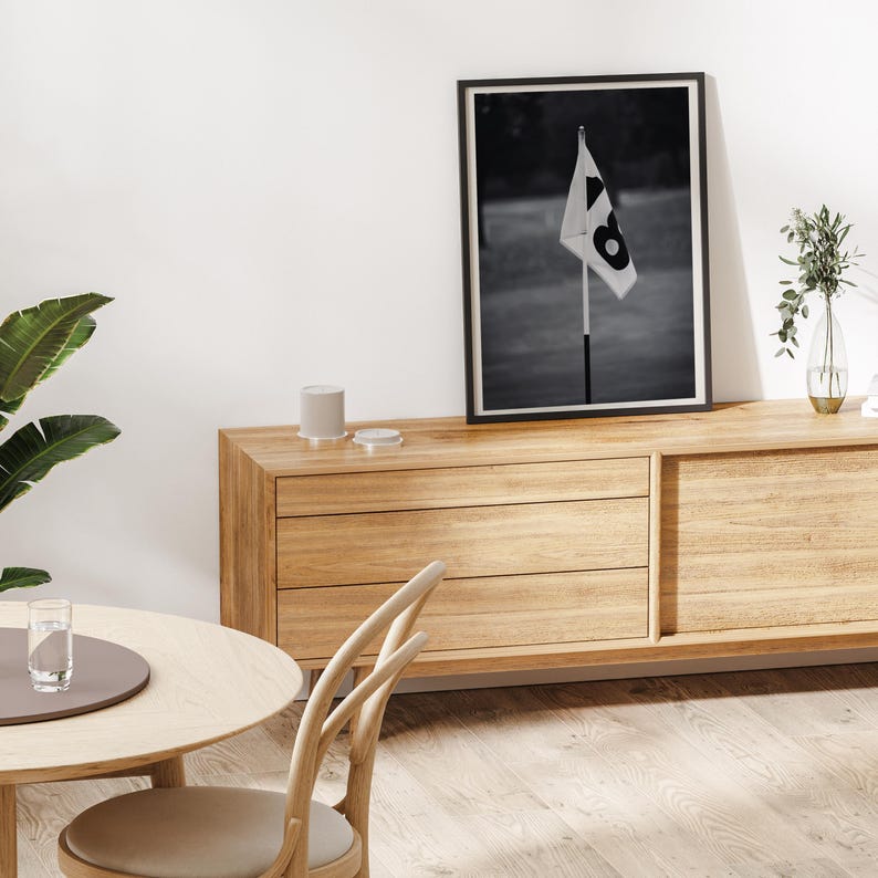 May include: Black and white framed print of a golf course flag with the number 8. The flag is white with a black number 8. The print is displayed on a light wood cabinet with a round table and chair in the foreground.