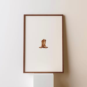 May include: A framed print featuring a pair of brown cowboy boots. The boots are centered on a white background, within a dark brown wooden frame. The artwork is displayed on a white pedestal against a neutral wall.