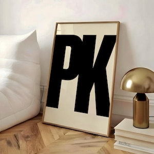 May include: A framed art print featuring the bold black letters "PK" against a cream background. The artwork is displayed in a room with a gold-colored lamp and a stack of books. The floor has a wooden parquet design.
