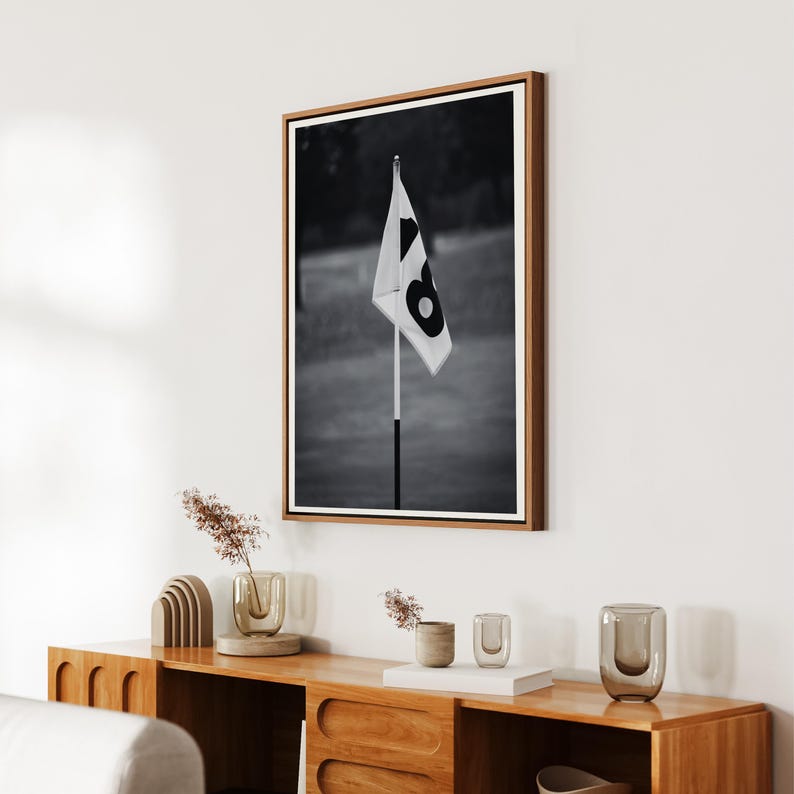 May include: Black and white framed photograph of a golf course flag. The flag is white with a large black number 8. The photograph is in a wooden frame and hangs on a white wall. The image is a close-up of the flag.