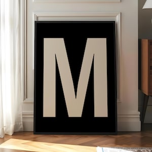 Op de afbeelding: Een ingelijste print met een grote, beige letter "M" op een zwarte achtergrond. Het kunstwerk wordt getoond in een kamer met een witte muur, houten vloer en een tapijt.