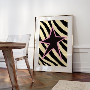 May include: Framed art print featuring a black star with a pink outline on a gold and black striped background. The artwork is displayed in a gold frame, leaning against a white wall. The print adds a modern touch to any room.