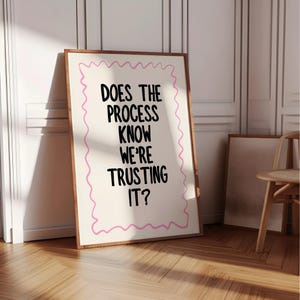 Op de afbeelding: Een ingelijste print met de tekst "DOES THE PROCESS KNOW WE'RE TRUSTING IT?" in zwart, tegen een crèmekleurige achtergrond met een roze golvende rand. De lijst is bruin en de print leunt tegen een witte muur.