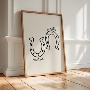 May include: A framed art print featuring two black horseshoe illustrations with dotted details. The words "in dallas" and "meet me" are handwritten in black script. The artwork is set against a cream background.