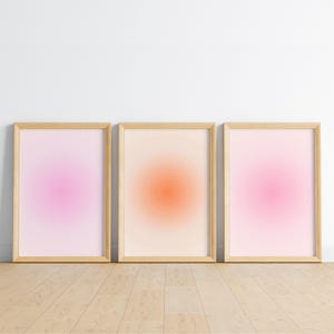 May include: Three framed art prints with a soft, gradient design. The prints feature a central, circular burst of color in shades of pink and orange, set against a light background. The frames are a natural wood color.
