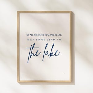 May include: A framed print with the text "OF ALL THE PATHS YOU TAKE IN LIFE, MAY SOME LEAD TO the lake" in navy blue script on a white background. The frame is a light wood color.