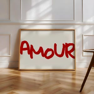 以下が含まれることがあります： クリーム色の背景に、赤色の太字で「AMOUR」と書かれた額入りのアートプリント。 アートワークは、壁の装飾に適した、明るい茶色の木製フレームに収められています。