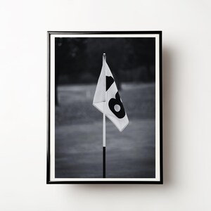 May include: Black and white framed photograph of a golf course flag. The flag is white with a large black number 6. The flag is on a pole, and the background is out of focus.