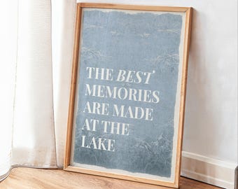 May include: A framed print with the text "THE BEST MEMORIES ARE MADE AT THE LAKE" in white letters on a light blue background. The wooden frame surrounds the print, which is leaning against a white wall and a curtain.