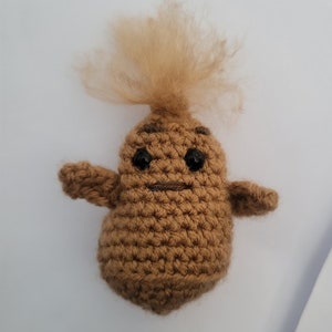Puede incluir: Un juguete de peluche de color marrón tejido a crochet con forma de patata y una peluca de pelo marrón esponjoso en la parte superior. El juguete tiene ojos negros y una boca cosida.