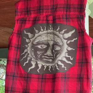 May include: Red and black plaid vest with a large brown patch featuring a sun face design. The vest is sleeveless and hangs on a blue hanger. The plaid pattern is visible on the back of the vest.