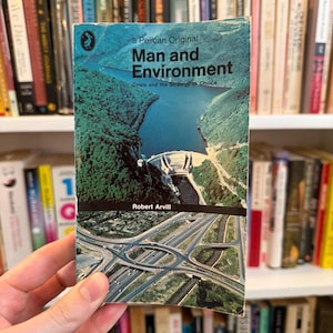Puede incluir: Un libro de bolsillo vintage titulado "Man and Environment" de Robert Arvill. La portada presenta una vista aérea escénica de una presa y un cruce de autopistas. El libro se sostiene frente a una estantería llena de varios libros.