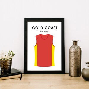 May include: A black framed print with a white background featuring a red sleeveless jersey with yellow side panels. The text "GOLD COAST EST 2009" is printed above the jersey.
