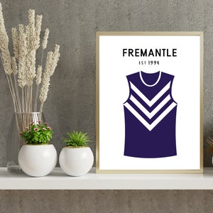 Puede incluir: Una impresión enmarcada con fondo blanco y un jersey sin mangas azul marino con rayas blancas en chevron. El texto "Fremantle Est 1994" está impreso en negro en la parte superior de la impresión.