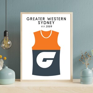 May include: A white framed print featuring a black and orange graphic of a sleeveless jersey with the letter "G" on the front. The text "GREATER WESTERN SYDNEY EST 2009" is at the top of the print.