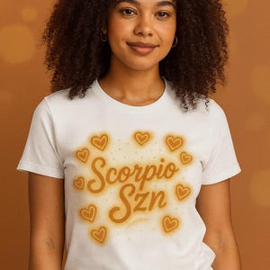 May include: White t-shirt with the text "Scorpio Szn" in a gold, glowing font, surrounded by gold heart shapes. The shirt is a crew neck style and is being worn by a person.