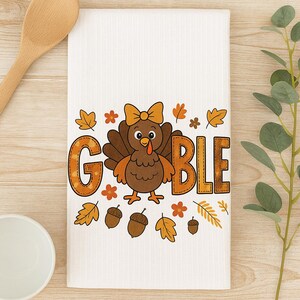 Könnte beinhalten: Weißes Küchentuch mit einem Cartoon-Truthahn-Design. Der Truthahn hat eine Schleife und ist von Herbstblättern, Eicheln und dem Wort "GOBBLE" in Orange und Braun umgeben.