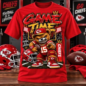 May include: A red short-sleeve t-shirt featuring a graphic of a brown teddy bear wearing a red cap, jersey with the number 15, and sneakers. The text reads GAME TIME, FOOTBALL IS A LIFESTYLE, and CHIEFS, with a football and helmet graphic.