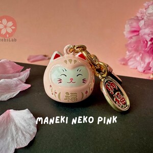 May include: Pink Maneki Neko charm with gold accents and a small gold charm with Japanese characters. The charm is on a black surface with pink flower petals.
