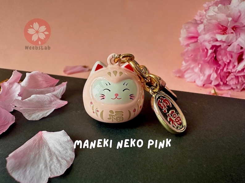 May include: Pink Maneki Neko charm with gold accents and a small gold charm with Japanese characters. The charm is on a black surface with pink flower petals.