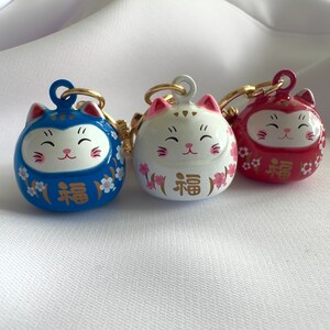 May include: Three lucky cat charms with gold accents. The charms are blue, white, and red with pink cherry blossoms and the word "福" (meaning "good fortune") in gold.